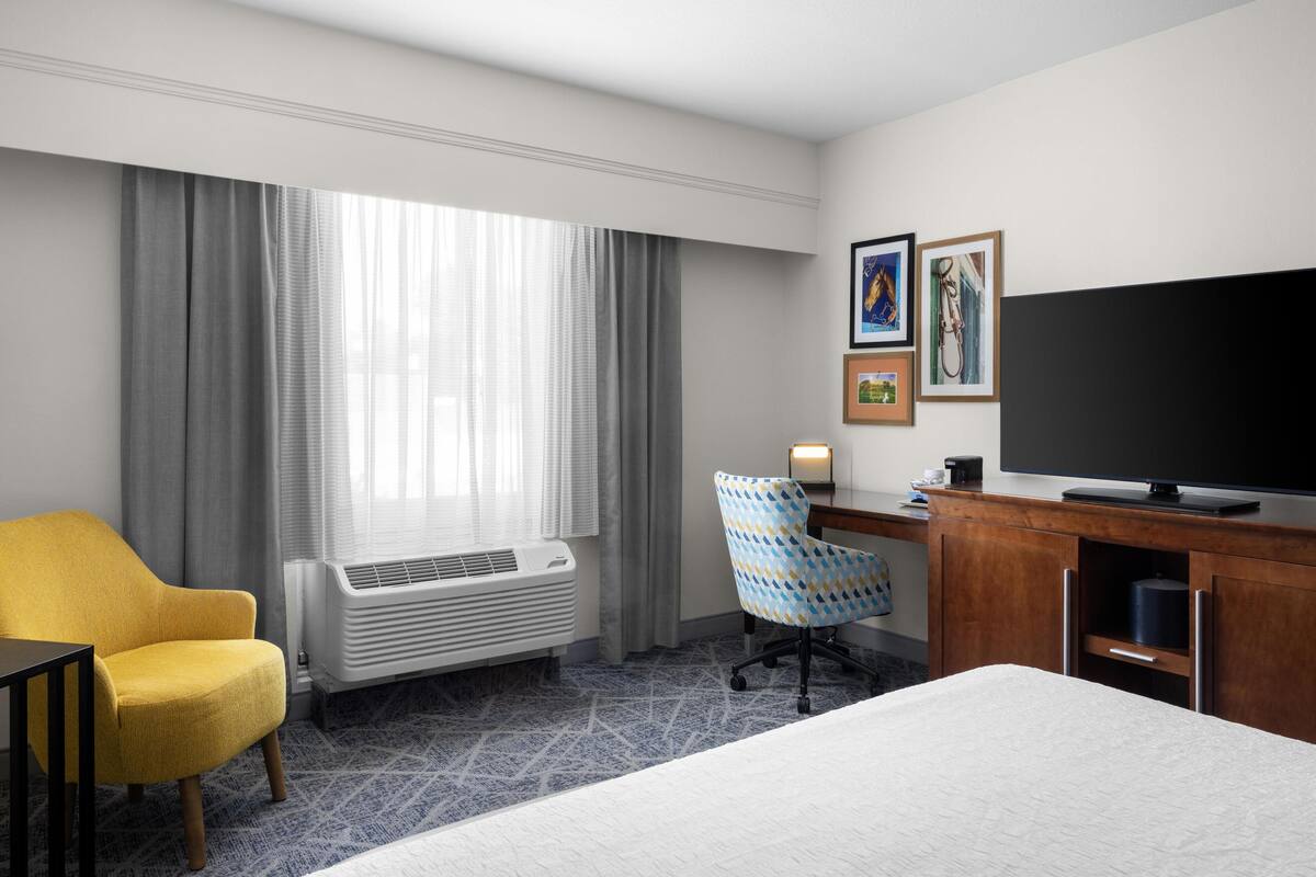 A hotel room with a bed, a yellow armchair, a desk with a TV, and a window with curtains.