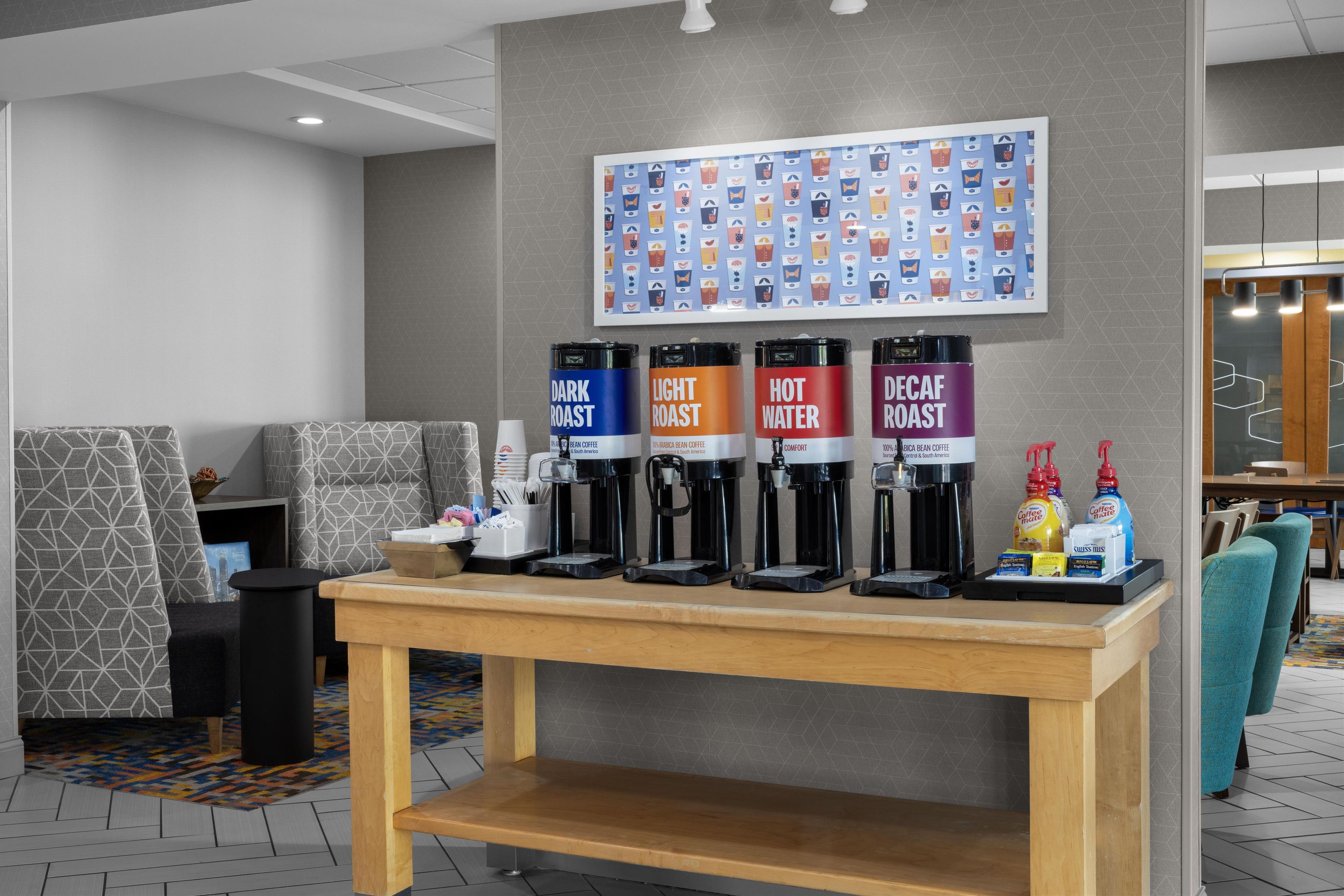 A coffee station with four coffee dispensers labeled 'Dark Roast,' 'Light Roast,' 'Hot Water,' and 'Decaf Roast.' There are condiments and a small table in front.