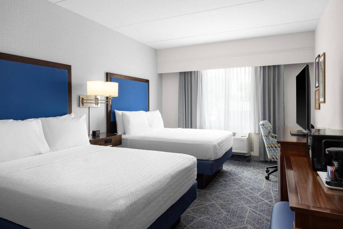 A hotel room with two beds, a desk, a chair, a TV, and a window with curtains.