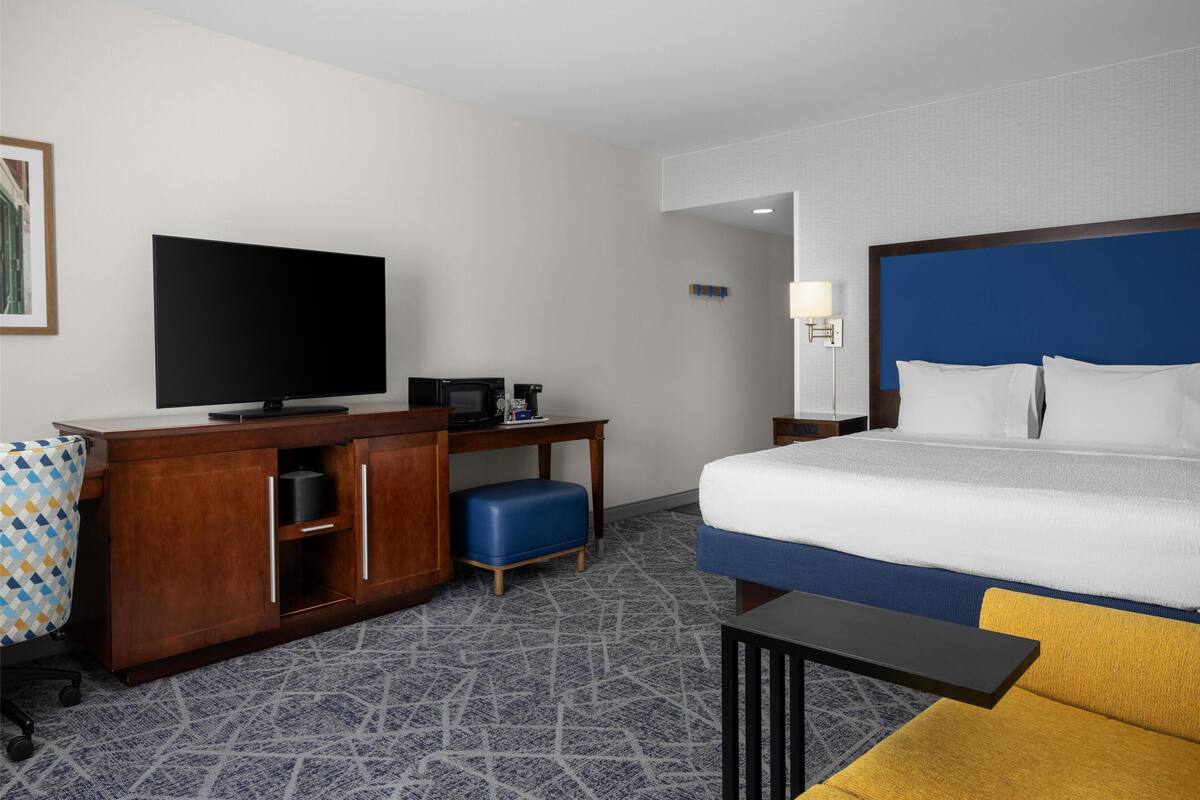 A hotel room with a large bed, a desk with a flat-screen TV, a coffee maker, and a blue ottoman.