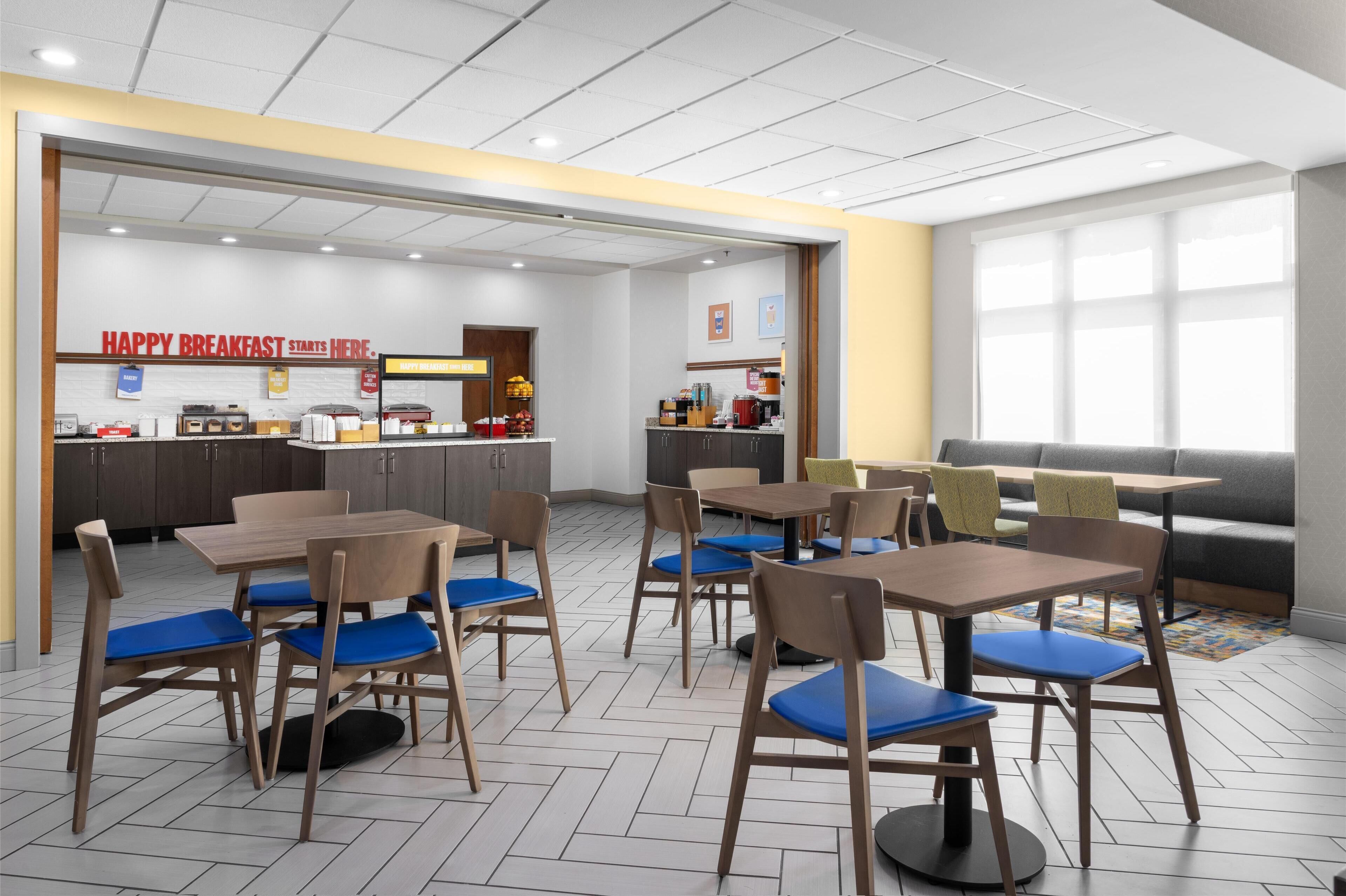A modern cafe with wooden tables and blue cushioned chairs, a kitchen area, and a seating area with a sofa and tables.
