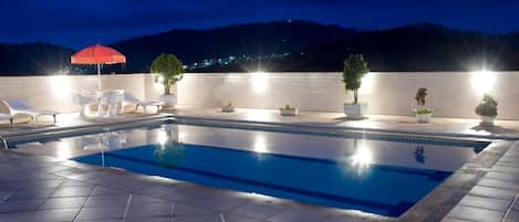 A swimming pool with tiled flooring, white lounge chairs, and potted plants.