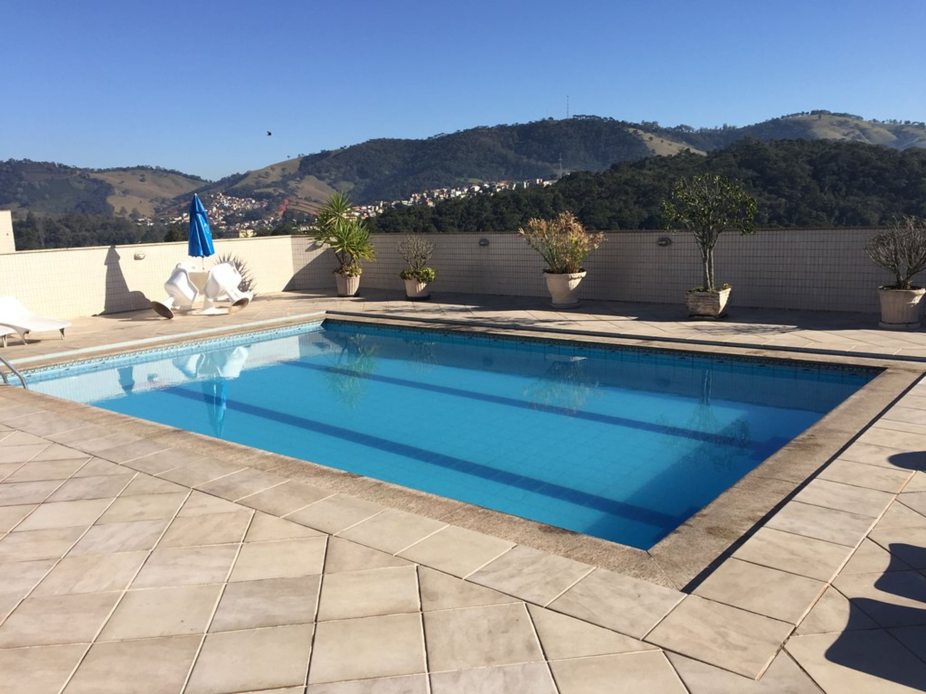Una piscina rectangular con bordes de azulejos, rodeada de plantas en macetas y sillas de descanso en una terraza en la azotea con vistas a un paisaje montañoso.
