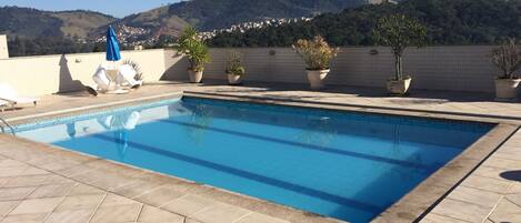 Una piscina rectangular con bordes de azulejos, rodeada de plantas en macetas y sillas de descanso en una terraza en la azotea con vistas a un paisaje montañoso.