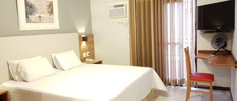 Ein Hotelzimmer mit einem großen Bett, einem Schreibtisch mit Stuhl, einem Fernseher und einem Fenster mit Vorhängen.