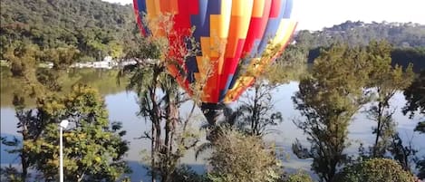 Un globo aerostático sobre un lago con árboles y un paisaje montañoso al fondo.
