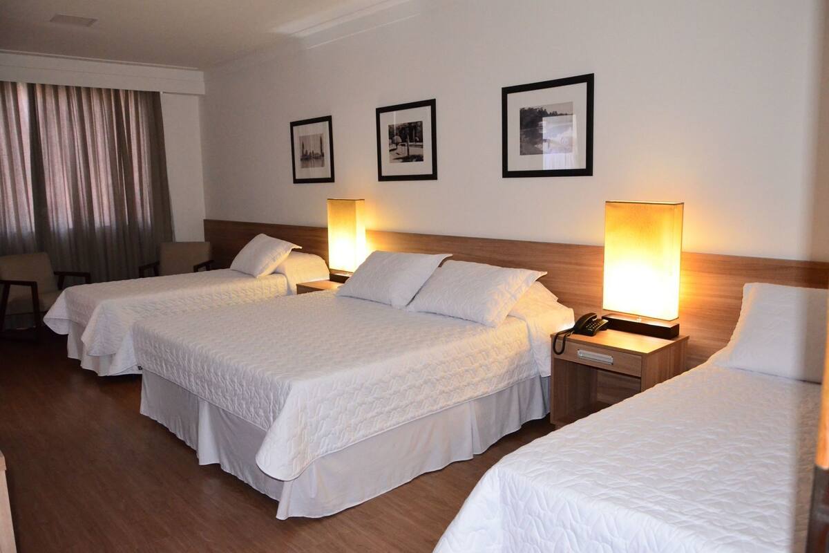 A hotel room with three beds, a desk, and a chair. There are three framed pictures on the wall.