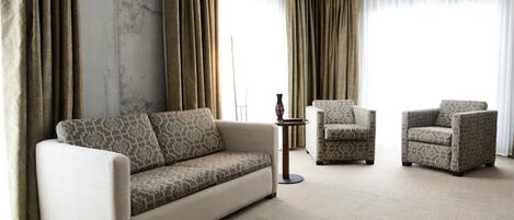 Un salón espacioso con un sofá y dos sillones, una mesita redonda y un amplio ventanal con cortinas.