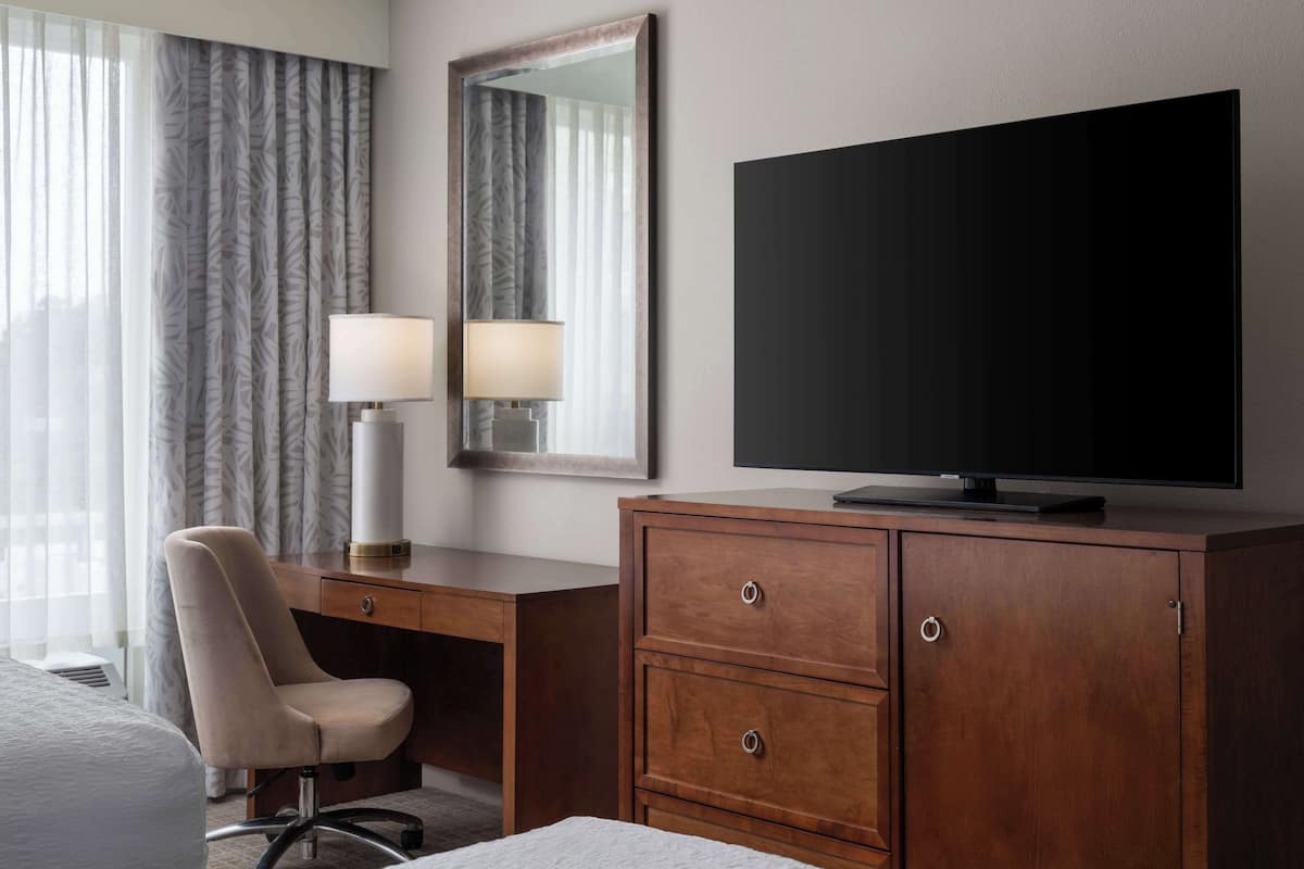 A hotel room with a wooden desk, a chair, a television, a mirror, and a window with curtains.