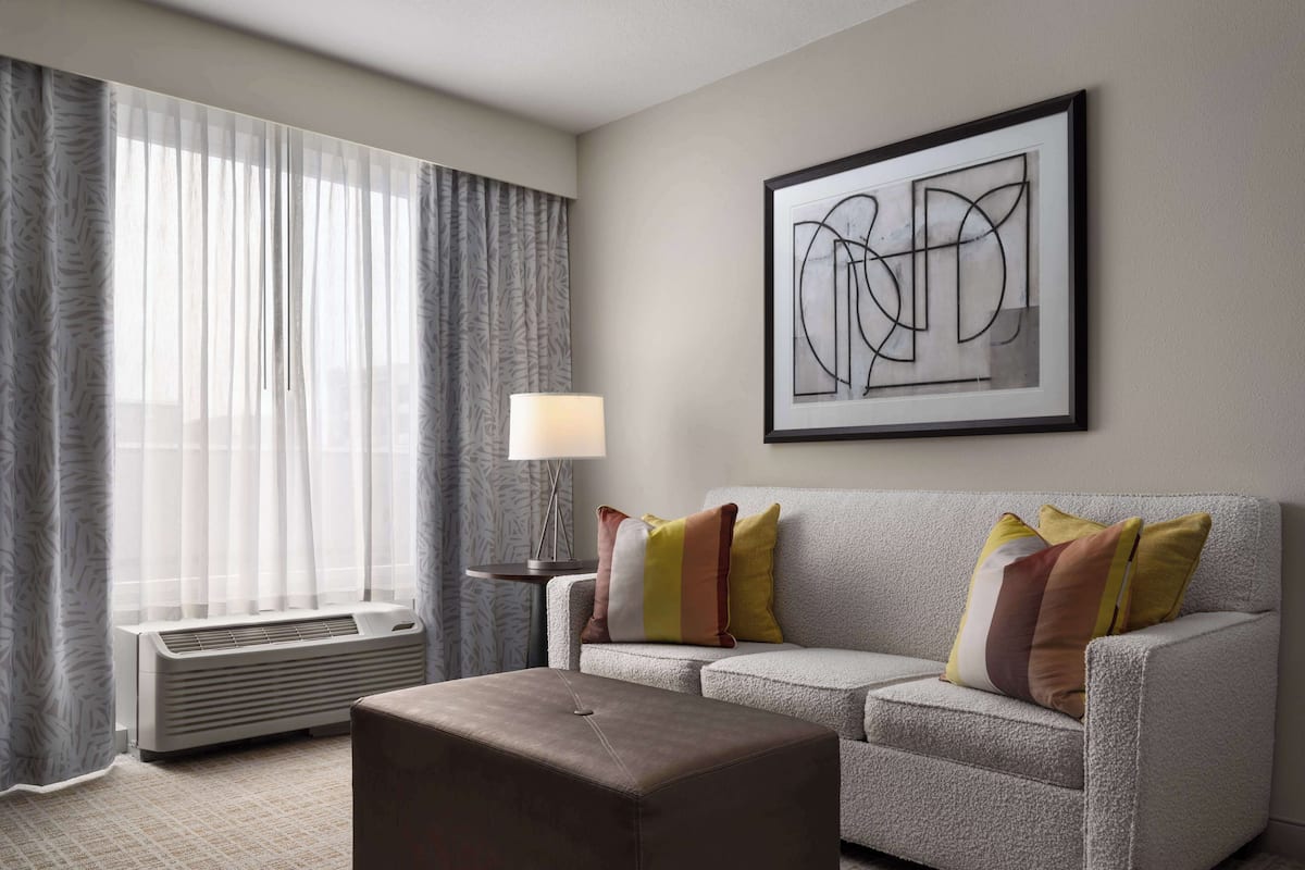 A modern hotel room with a grey sofa, a brown ottoman, a standing lamp, and a large abstract painting on the wall.