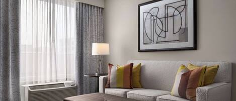 A modern hotel room with a grey sofa, a brown ottoman, a standing lamp, and a large abstract painting on the wall.