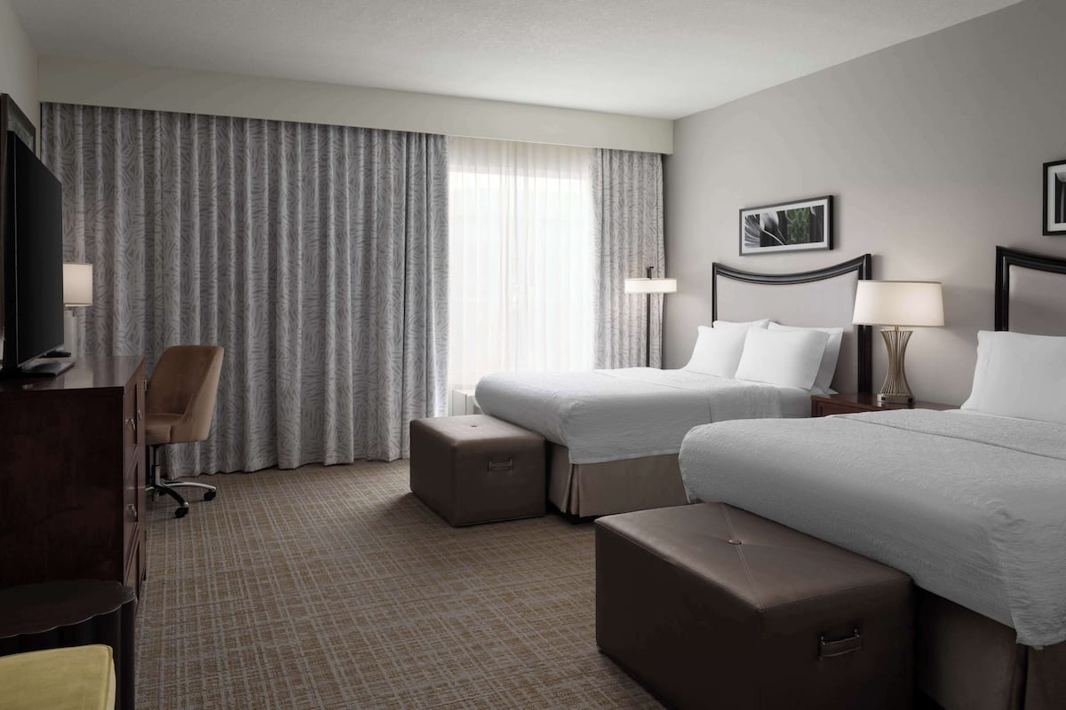 A hotel room with two beds, a desk, a chair, and a large window with curtains.