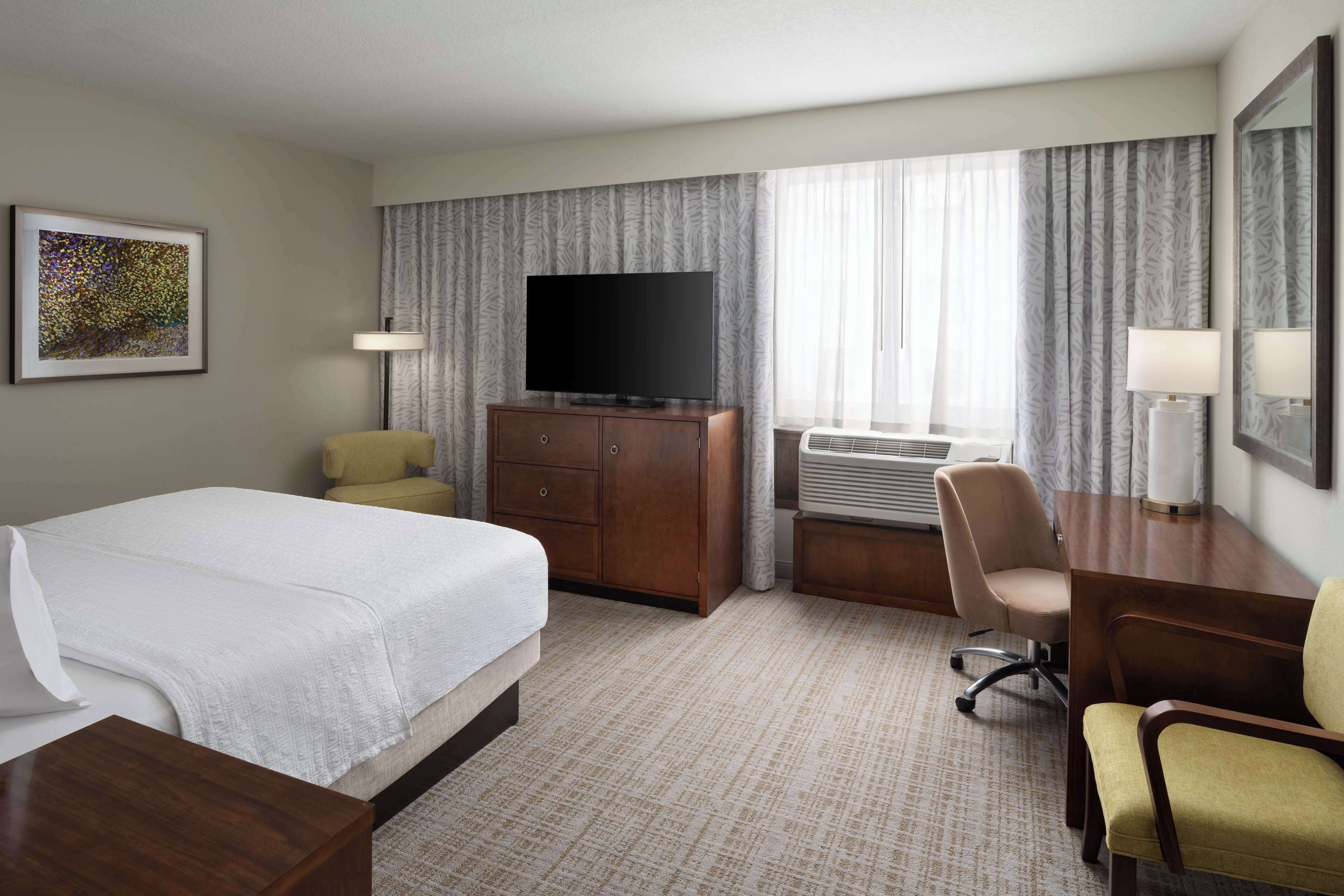 A hotel room with a bed, a desk, a chair, a television, and an air conditioning unit.