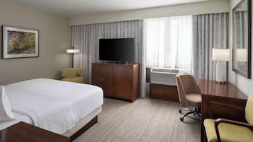 A hotel room with a bed, a desk, a chair, a television, and an air conditioning unit.