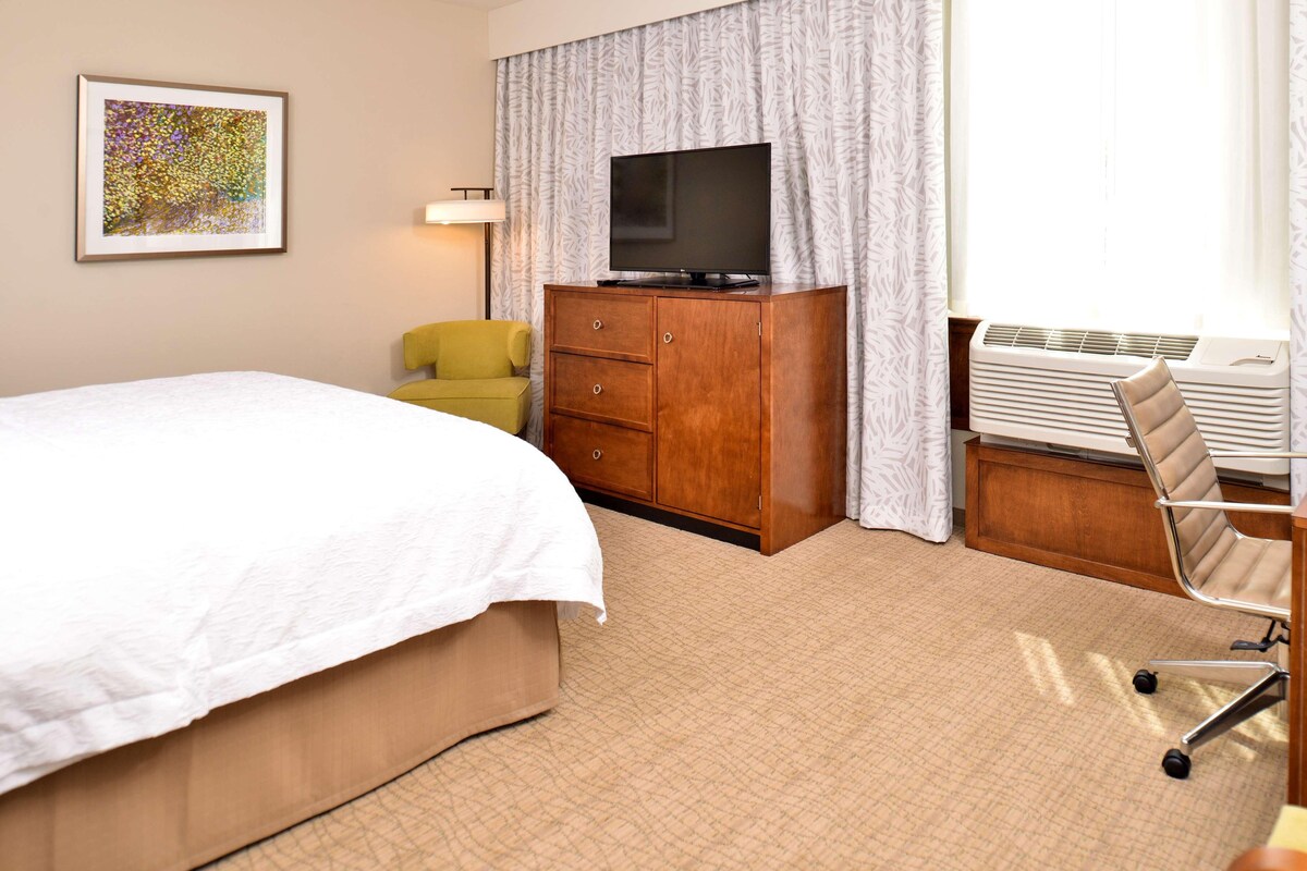 A hotel room with a bed, a TV on a wooden cabinet, a desk with a chair, and a window with curtains.