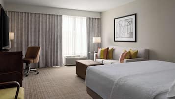 Habitación de hotel con una cama grande, un escritorio, una silla, un sofá y un ventanal con cortinas.
