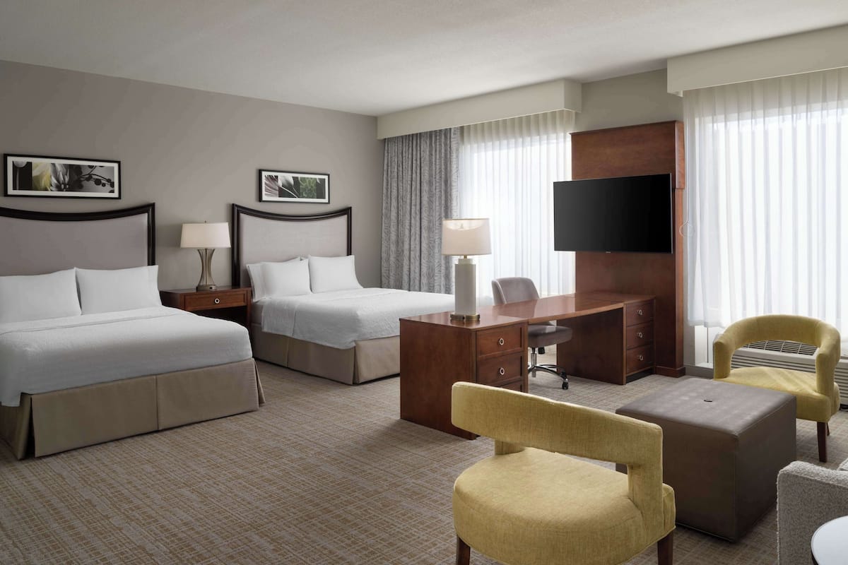 A hotel room with two beds, a desk, a chair, a TV, and a large window with curtains.