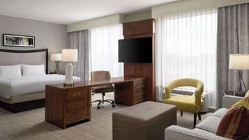 A hotel room with a bed, desk, chair, ottoman, and a television.