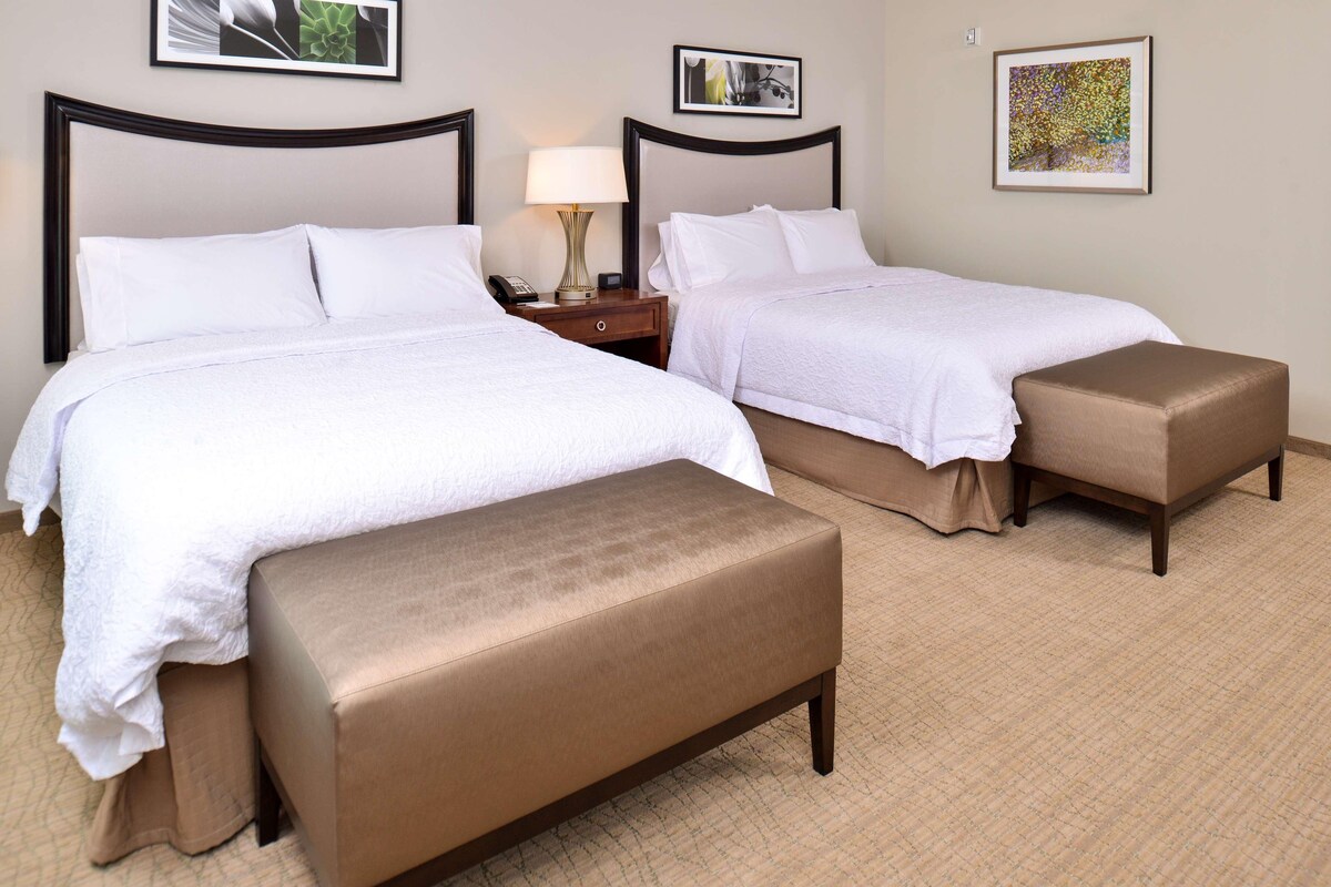 A hotel room with two beds, a nightstand with a lamp, and a brown bench.