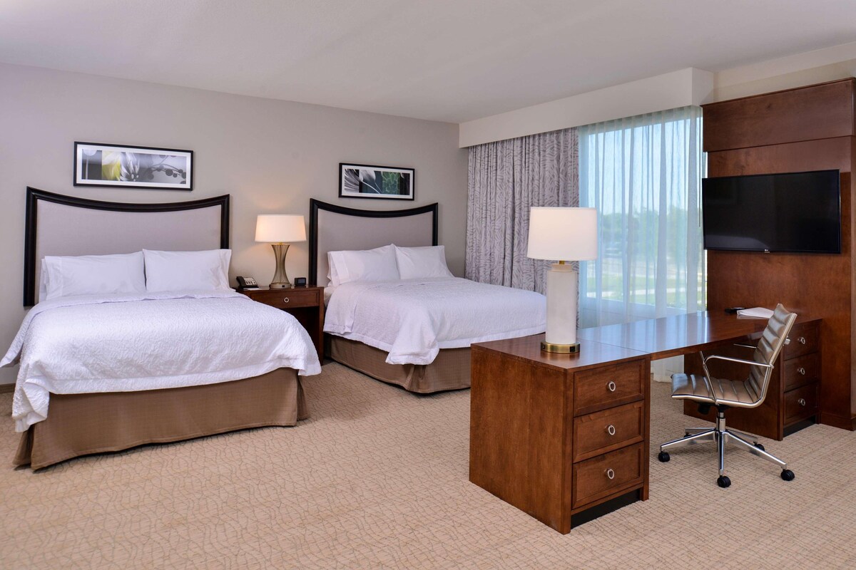A hotel room with two beds, a desk with a chair, a TV, and artwork on the walls.