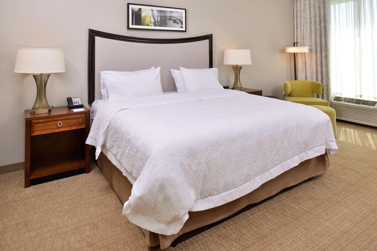 A hotel room with a large bed, two bedside tables with lamps, a chair, and a window with curtains.