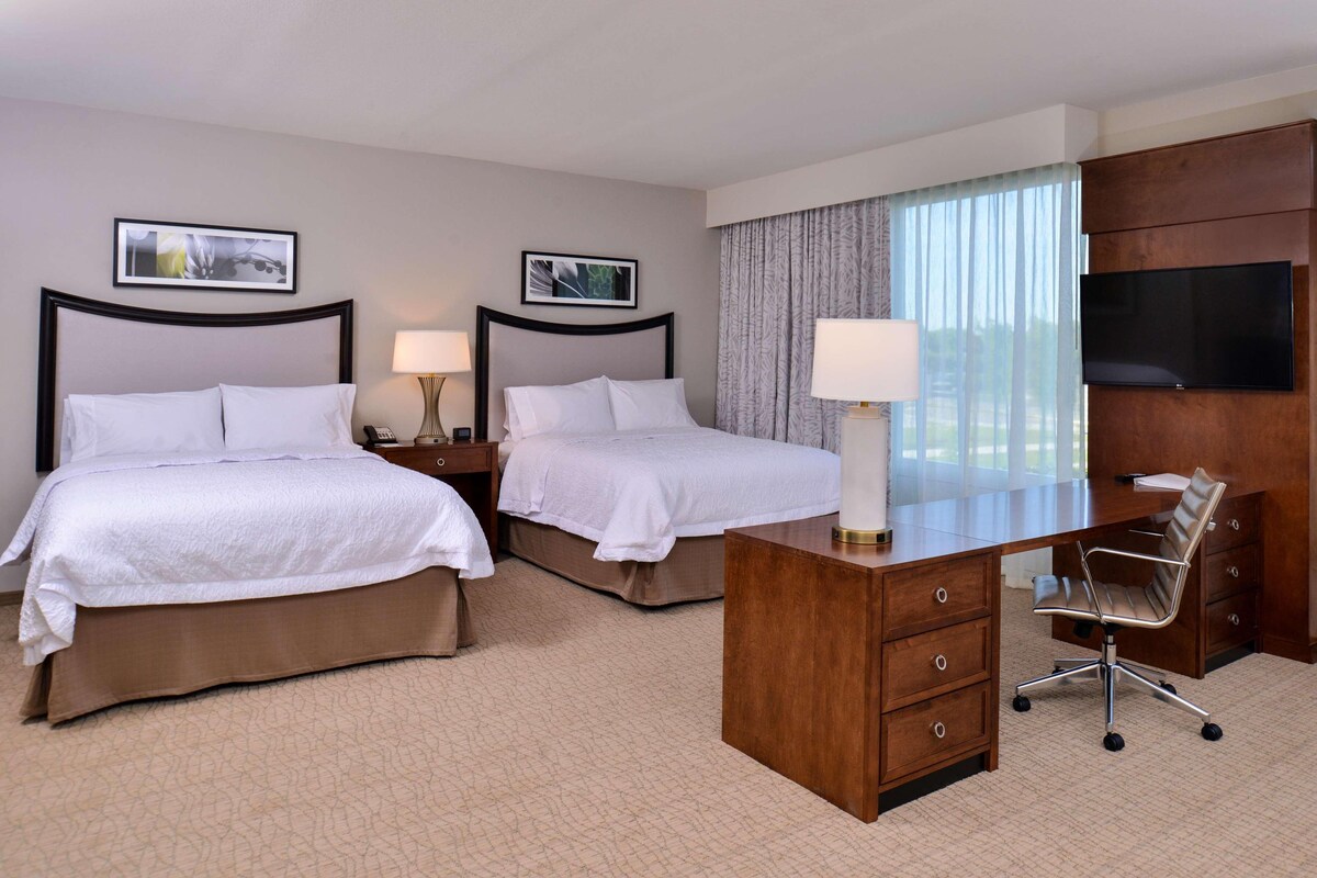 A hotel room with two beds, a desk, a chair, a TV, and a window with curtains.