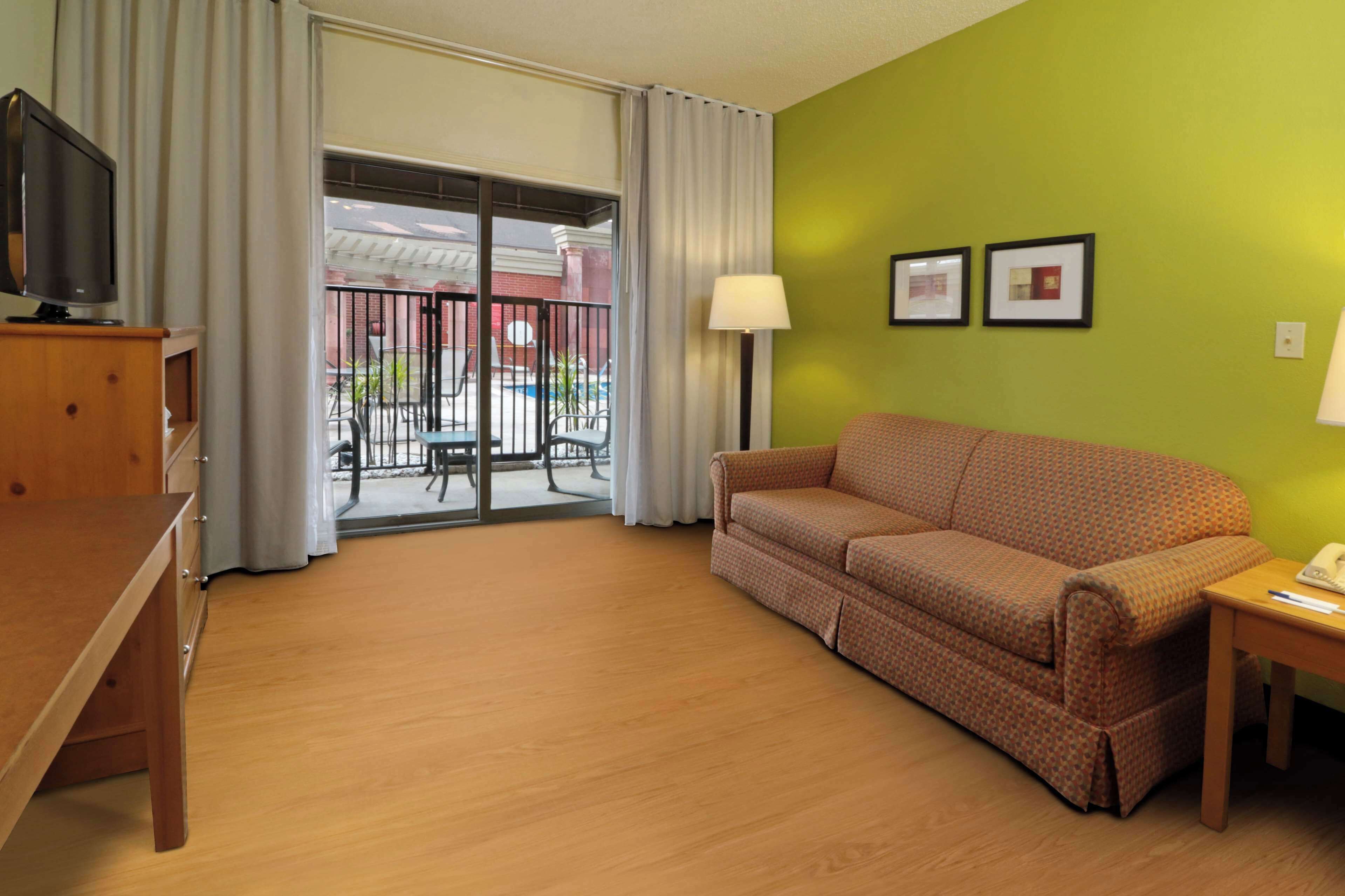 A hotel room with a sofa, a TV, a lamp, and a view of a pool and building through a glass door.