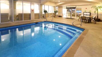An indoor swimming pool with clear glass walls, a tiled floor, and a seating area with tables and chairs.
