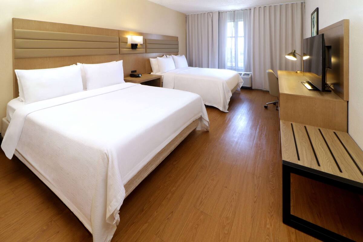 A hotel room with two beds, a desk, a TV, and a window with curtains.