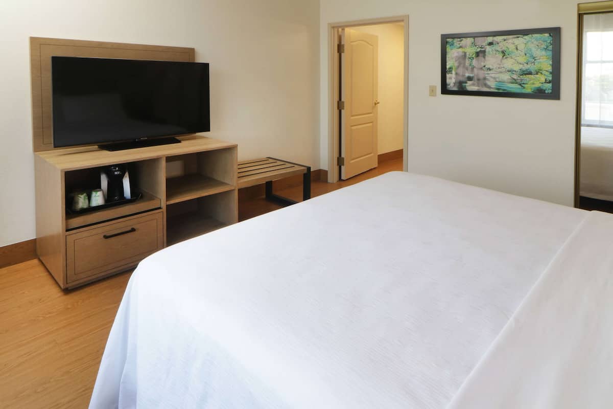 A hotel room with a bed, a TV on a wooden stand, a painting on the wall, and a door leading to another room.
