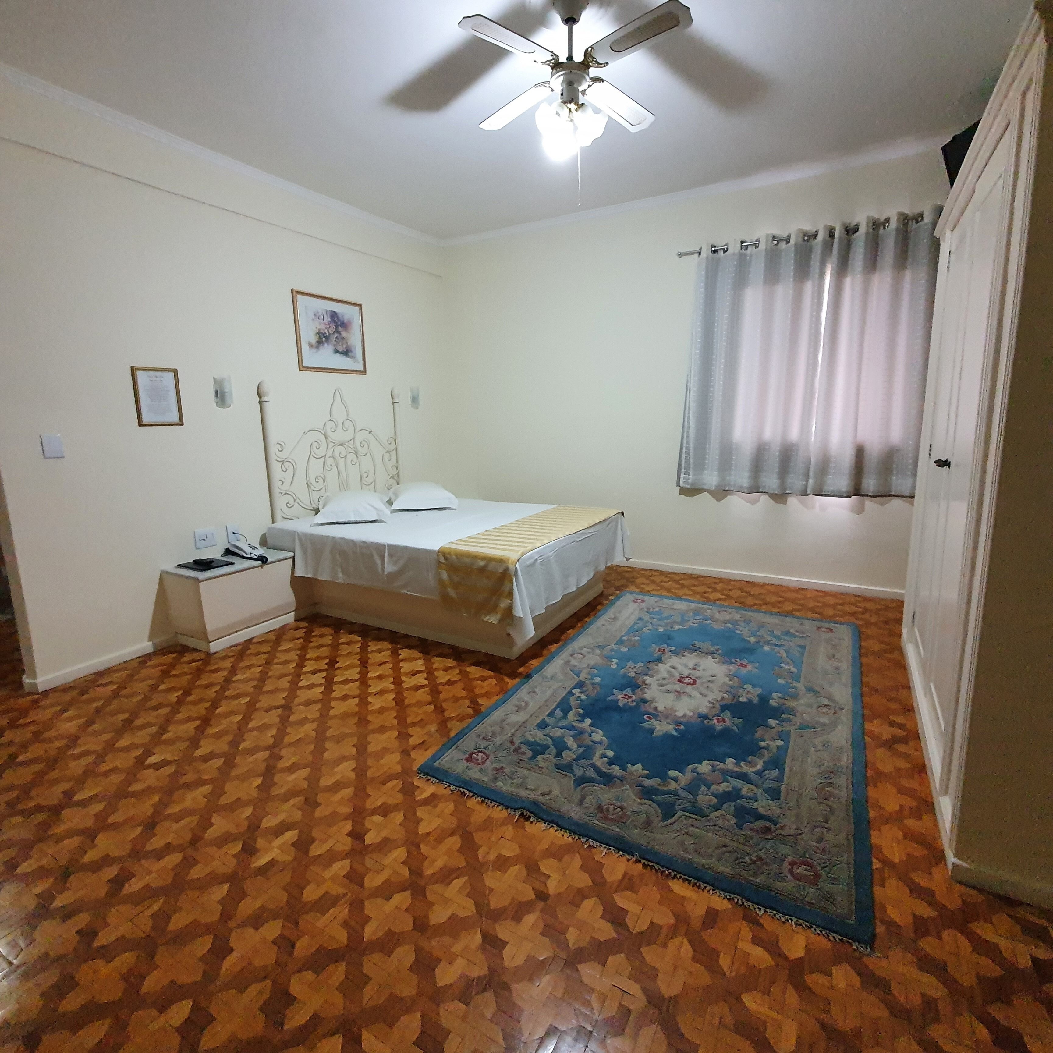 A bedroom with a bed, a nightstand, a ceiling fan, a window with curtains, and a patterned rug.