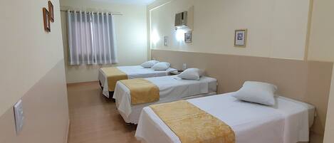 Habitación de hotel con tres camas individuales, cada una con cubrecama blanco y un plaid amarillo. Hay una ventana con cortinas blancas, una lámpara de techo y un equipo de aire acondicionado instalado en la pared.