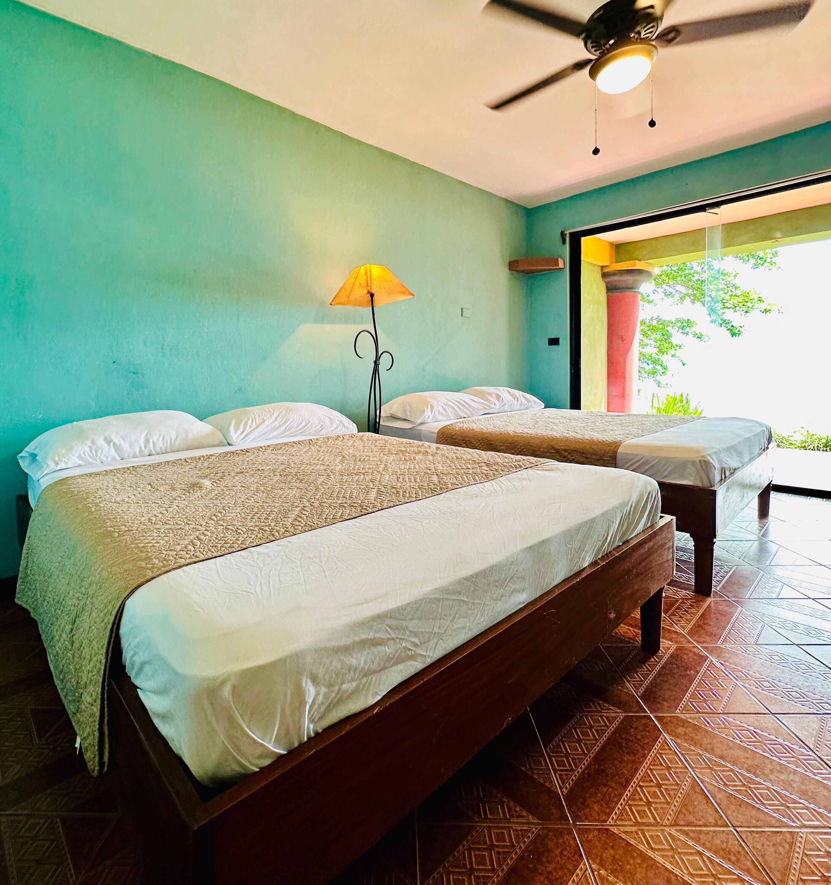 A hotel room with two beds, a ceiling fan, and a view of greenery outside.