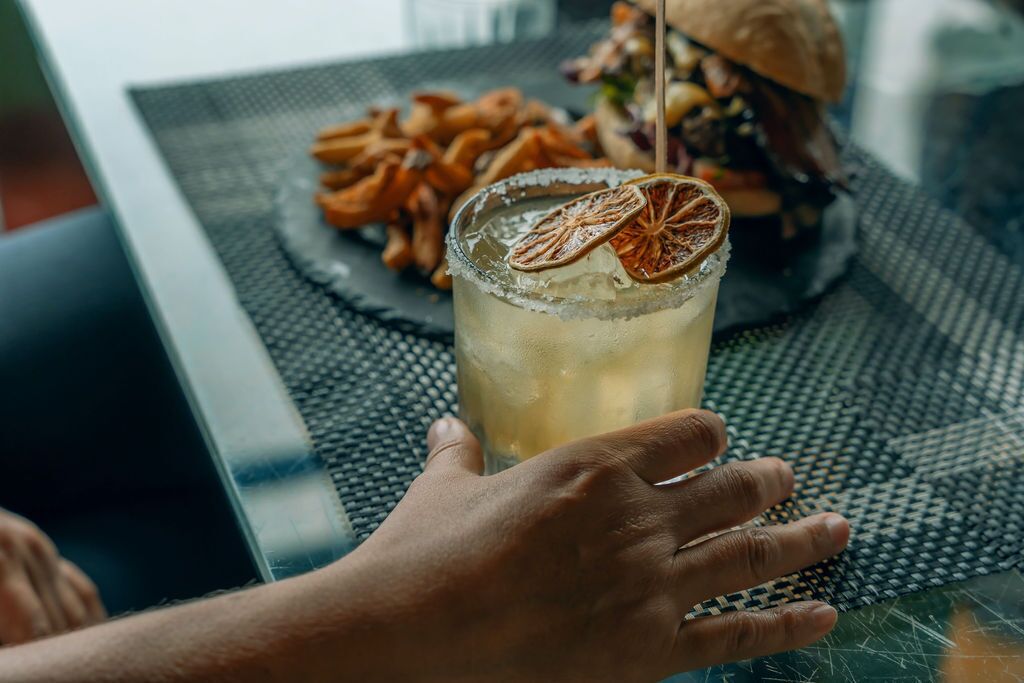 A hand holding a glass with a beverage, a plate of food, and a burger on a table.