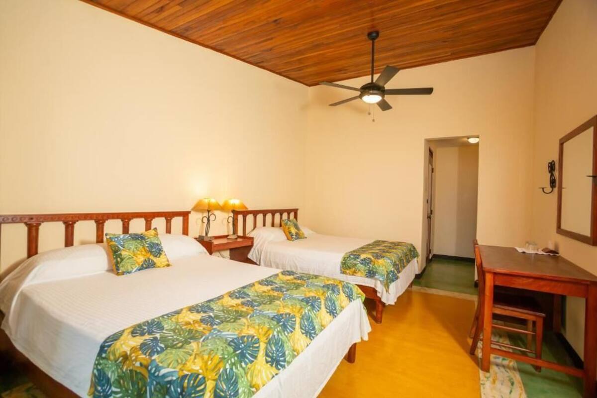 A room with two beds, a ceiling fan, a desk, and a door leading to another room.