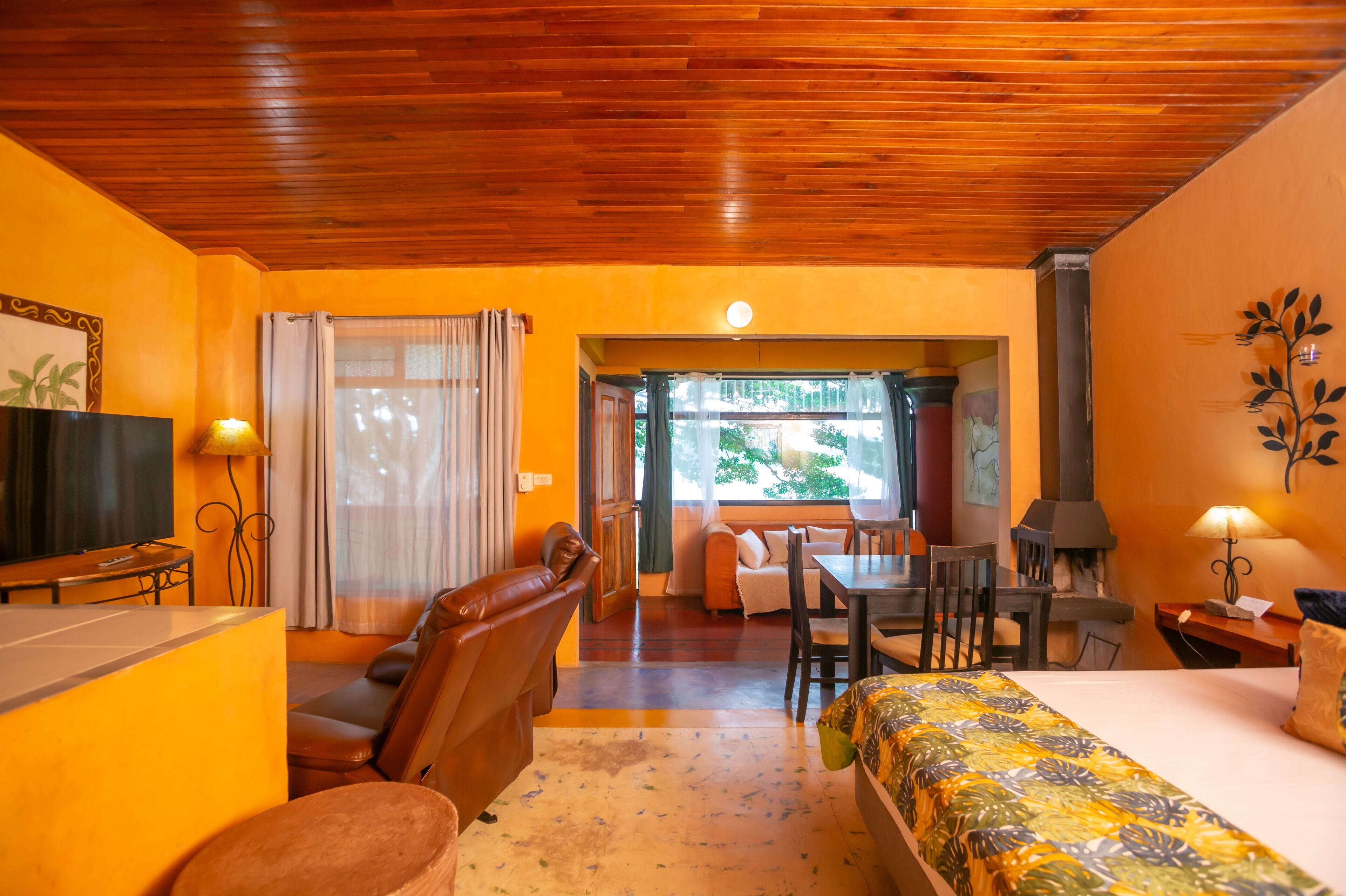 A hotel room with a wooden ceiling, a bed with a floral bedspread, a TV, a sofa, a dining table with chairs, and a fireplace.
