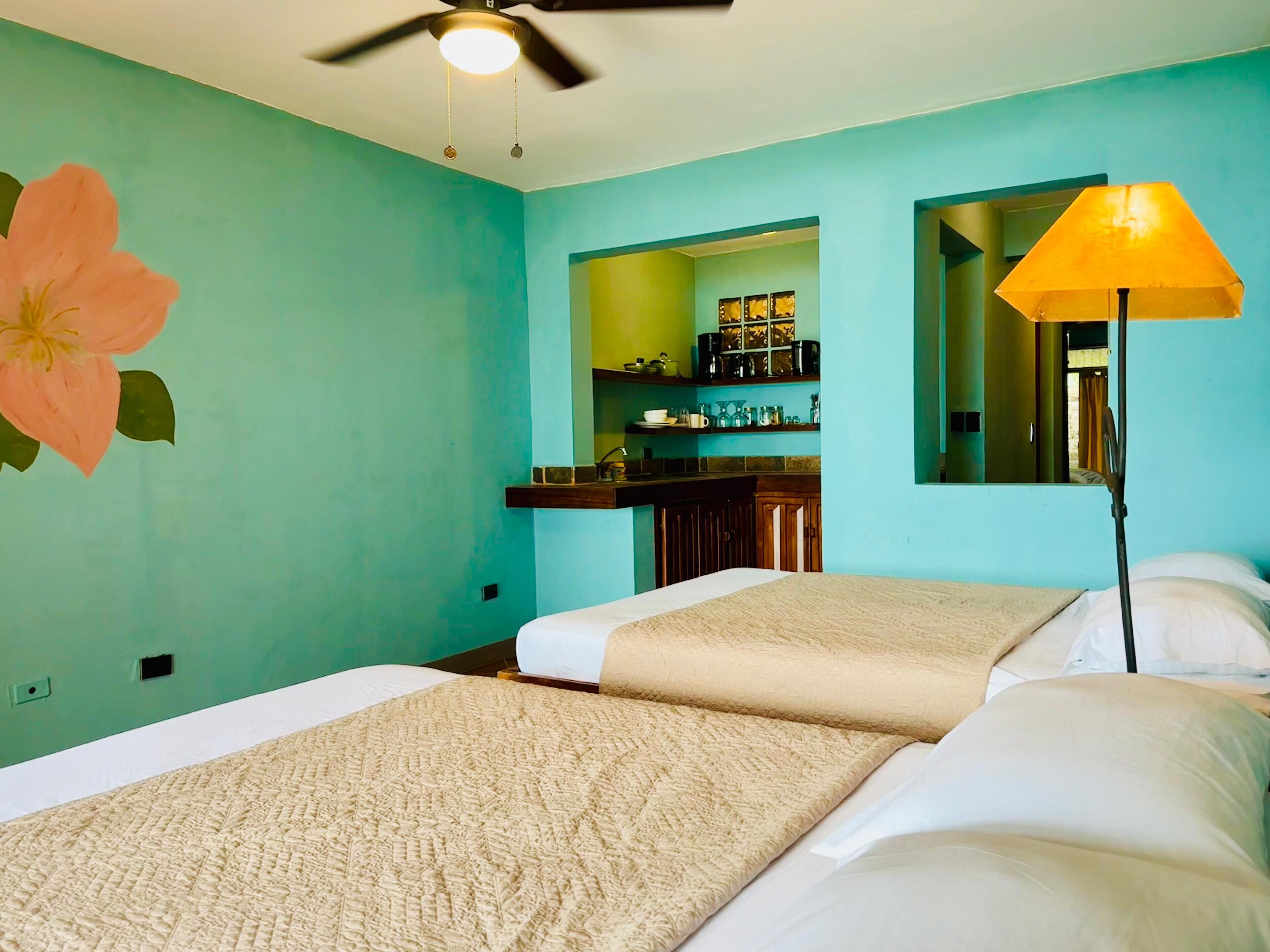 Habitación de hotel con dos camas, ventilador de techo, un gran mural de flores, zona de cocina con estantes y una lámpara de pie.
