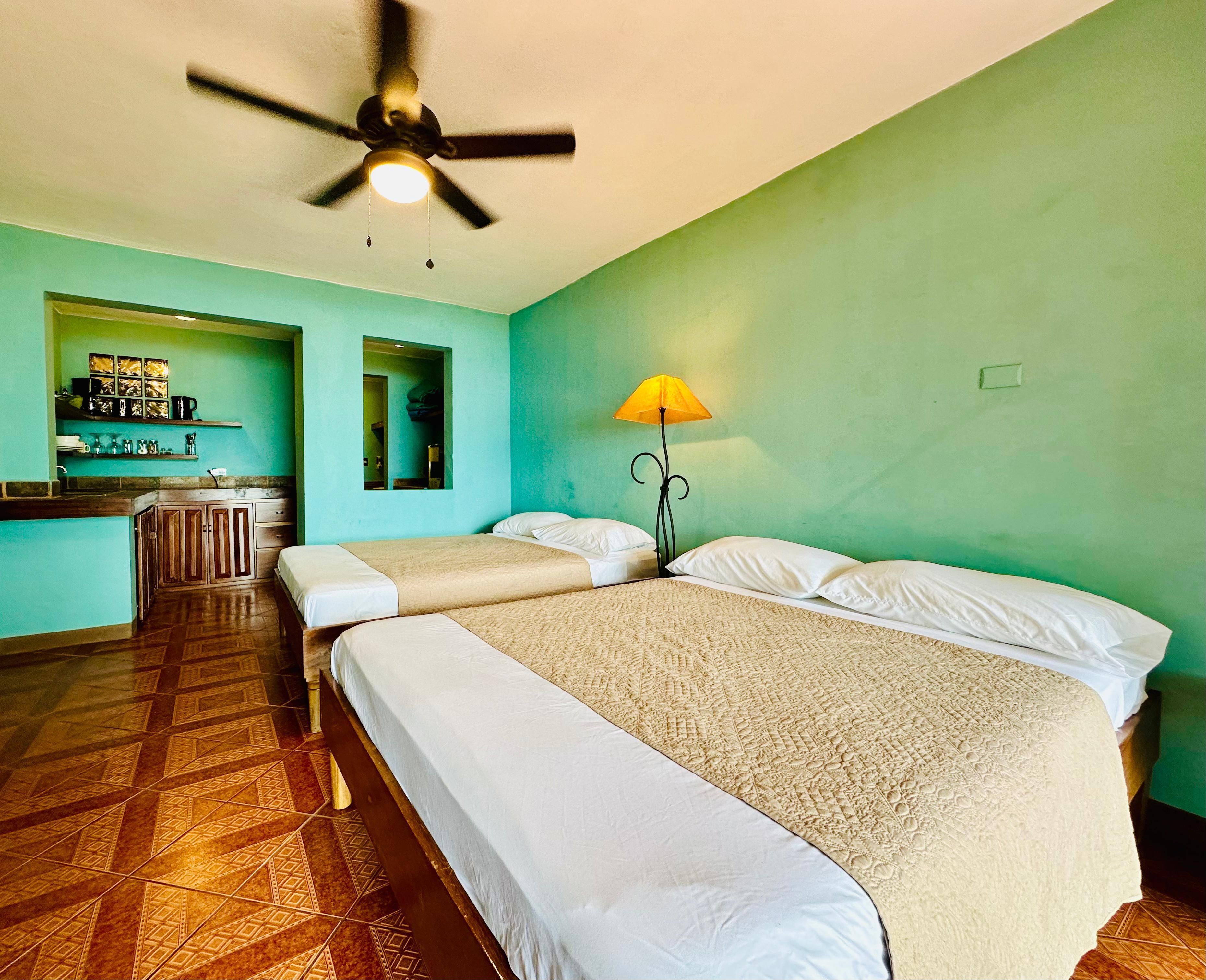 A room with two beds, a ceiling fan, a small kitchen area, and a green wall.