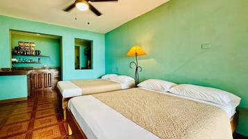 A room with two beds, a ceiling fan, a small kitchen area, and a green wall.