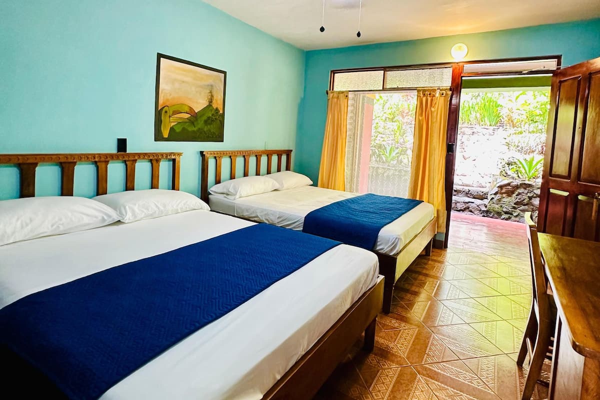 A room with two beds, a ceiling fan, a painting, and a view of greenery outside.