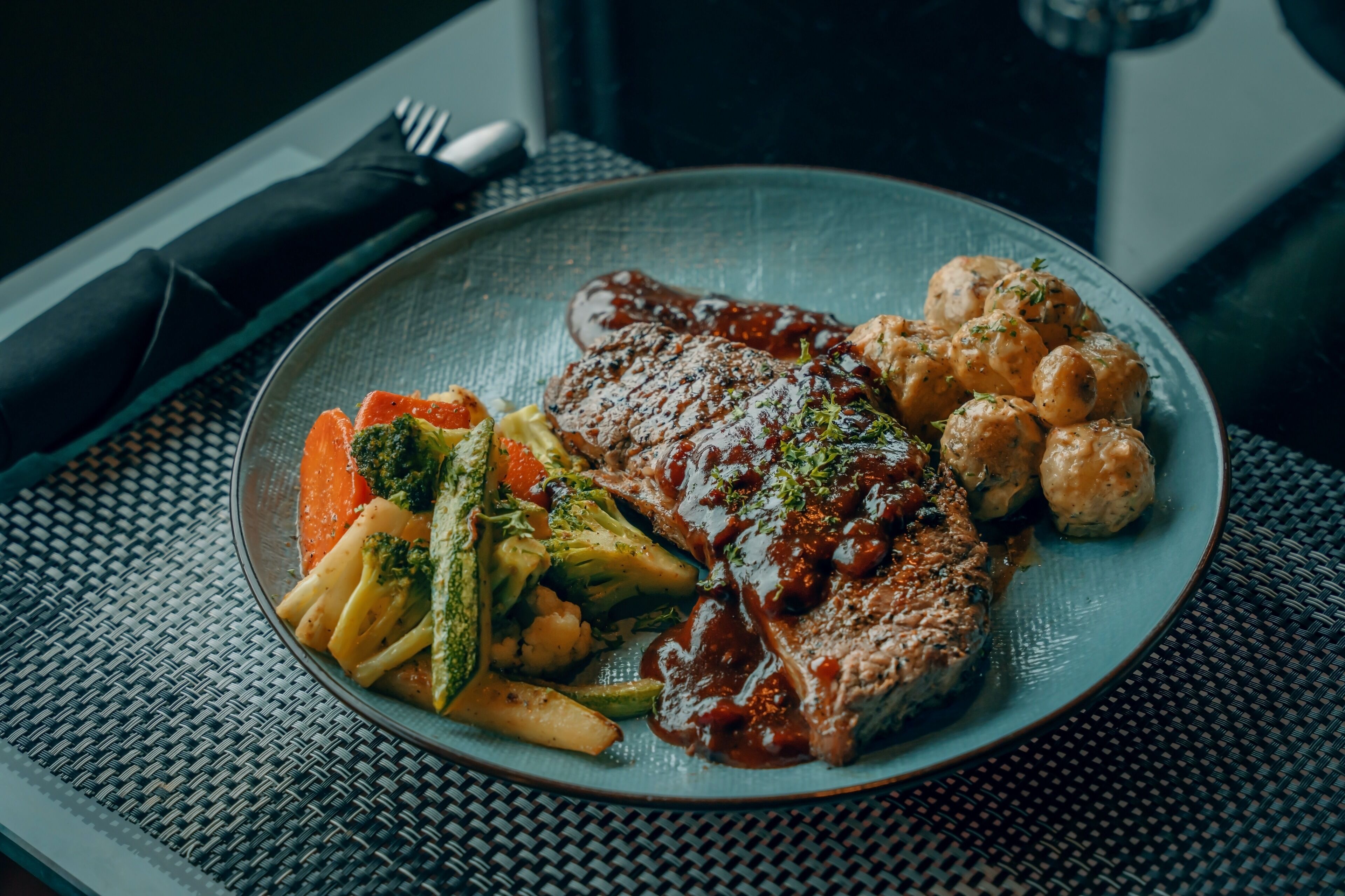 Un plato de comida con un filete, verduras y dumplings sobre un mantel tejido.