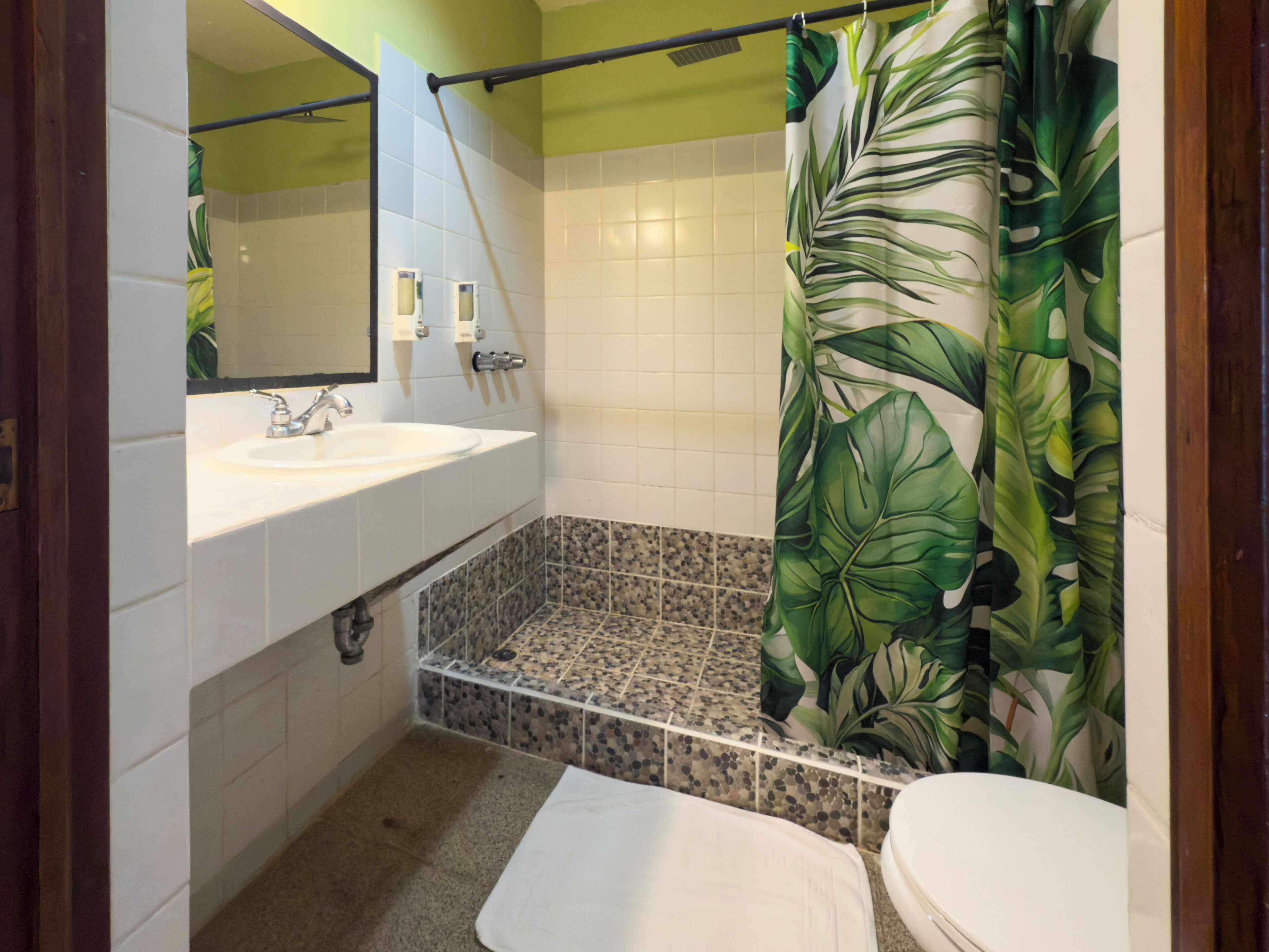 Un bagno con un lavandino bianco, uno specchio, una doccia con tenda a motivi fogliame e pavimento piastrellato.