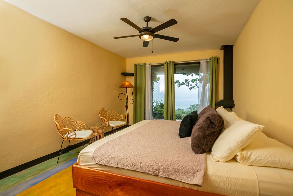 A bedroom with a bed, a ceiling fan, a window with curtains, and a view of greenery outside.