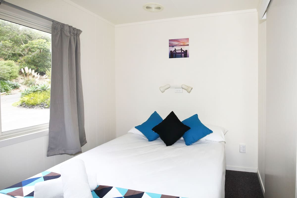 A neatly made bed with a geometric-patterned bedspread, a window with a view of greenery, and a small picture on the wall.