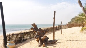 Une plage de sable bordée d’une clôture en bois, avec des palmiers et une structure rustique également en bois.