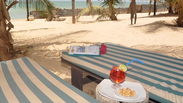 Un salon de plage avec des coussins à rayures, un verre d’une boisson rouge agrémentée d’une tranche de citron vert, et un menu posé sur une petite table.