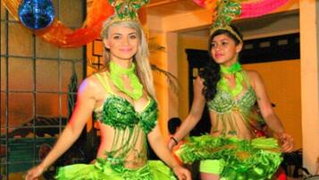 Two individuals in green costumes with feathered headdresses, standing in a room with a tiled wall and a door in the background.