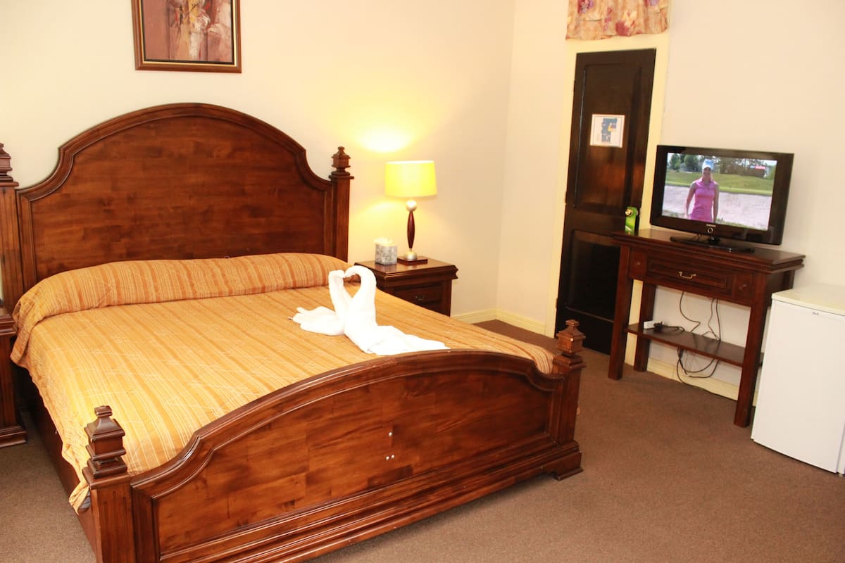 Een net opgemaakt bed met een houten hoofdbord, een bedlampje, een televisie op een standaard en een kleine witte koelkast.