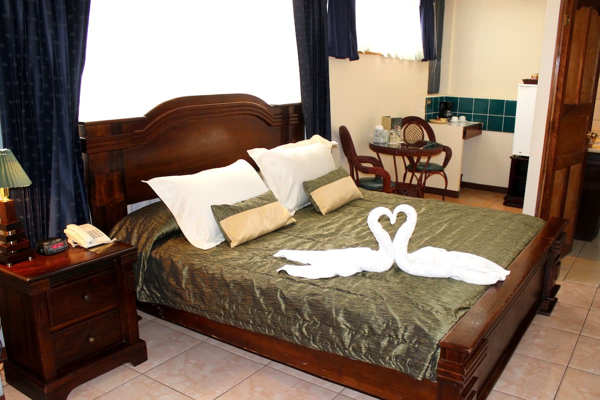 Een netjes opgemaakt bed met een groene sprei en twee zwaanvormige handdoeken, een houten nachtkastje met een lamp, en een stoel in een klein zithoekje.