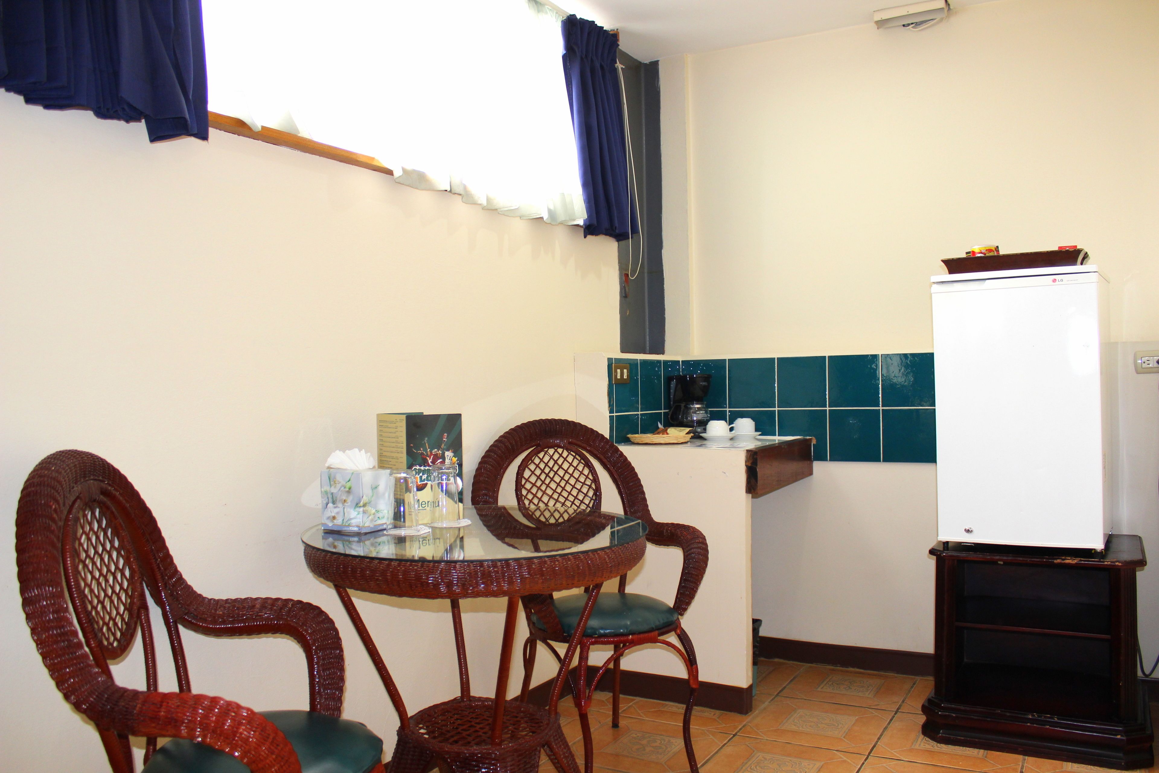 A small kitchen with a round table and two wicker chairs, a white refrigerator, and a tiled backsplash.
